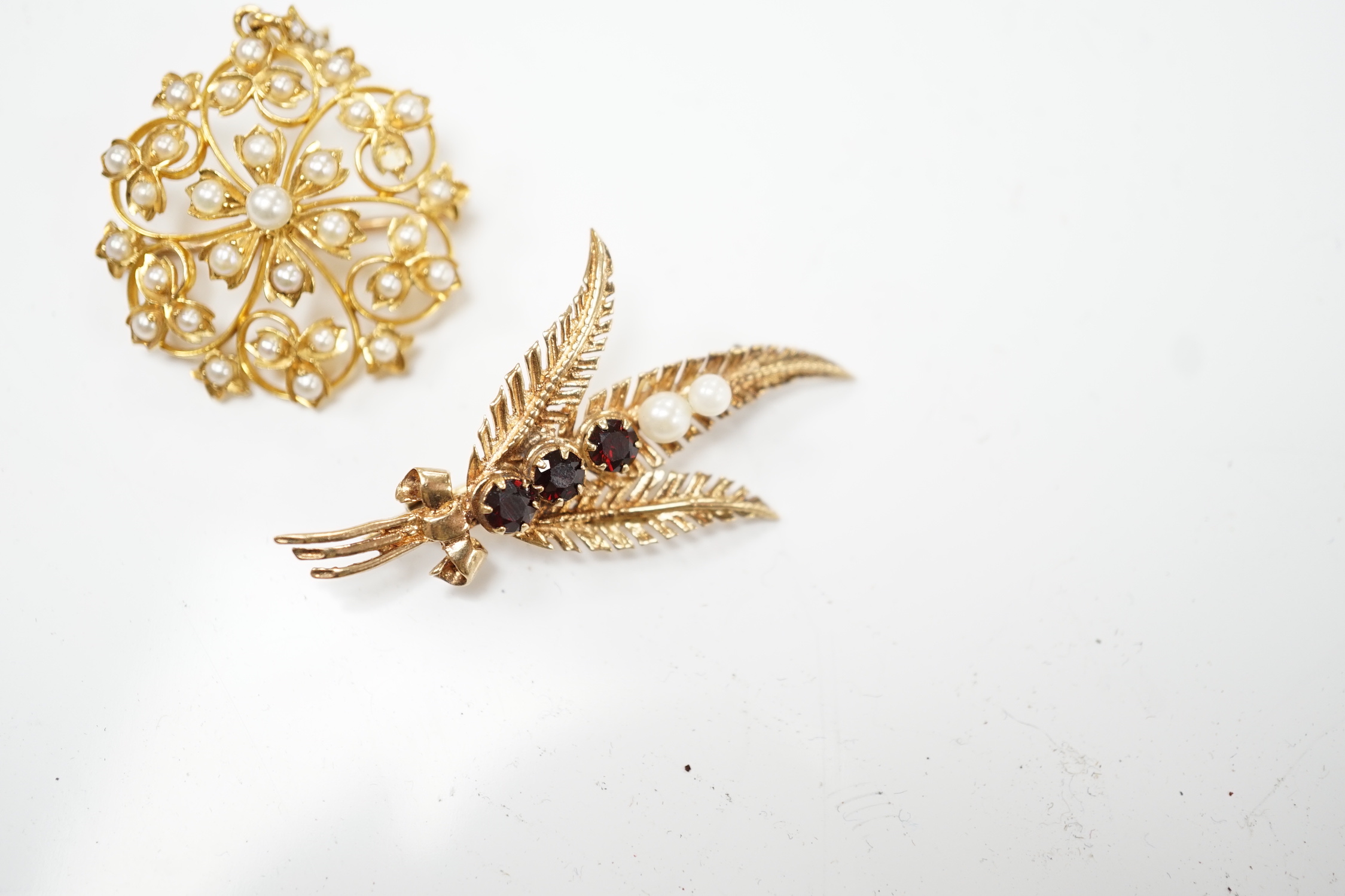An Edwardian yellow metal and seed pearl set circular cluster pendant brooch, 27mm, gross weight 6.2 grams, together with a later 9ct gold, garnet and cultured pearl set foliate brooch, gross weight 4.1 grams.